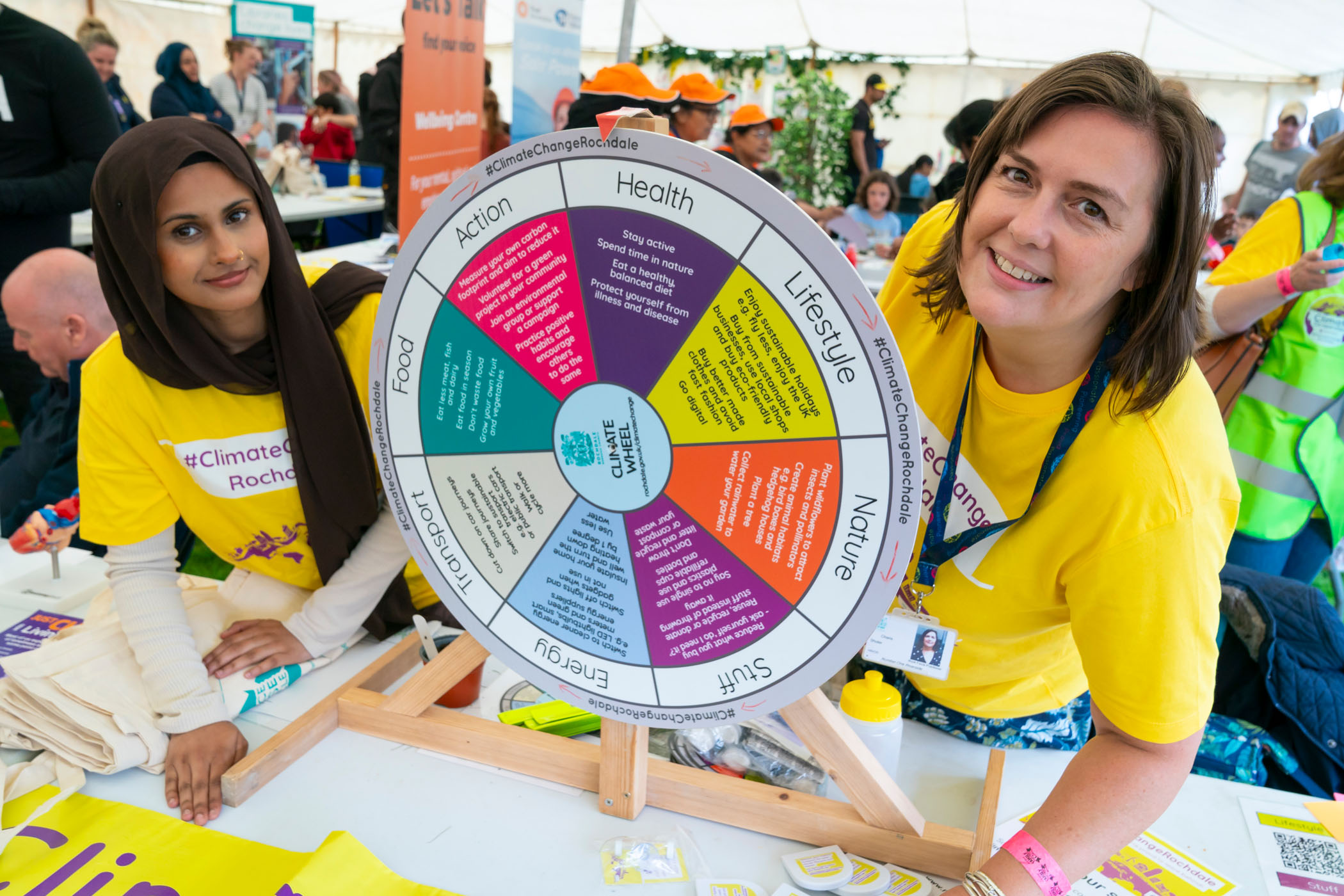 Two members of the #ClimateChangeRochdale team smile at at event alongside a wheel that can be spun to choose from suggested climate actions we can all take